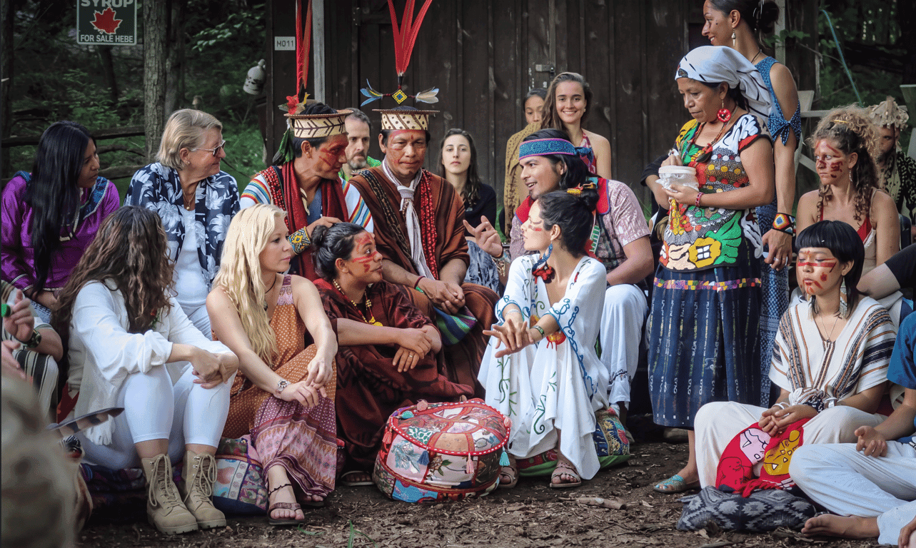 Indigenous elders and community members gathered in a circle outdoors, some in traditional ceremonial dress, engaged in conversation in a wooded setting