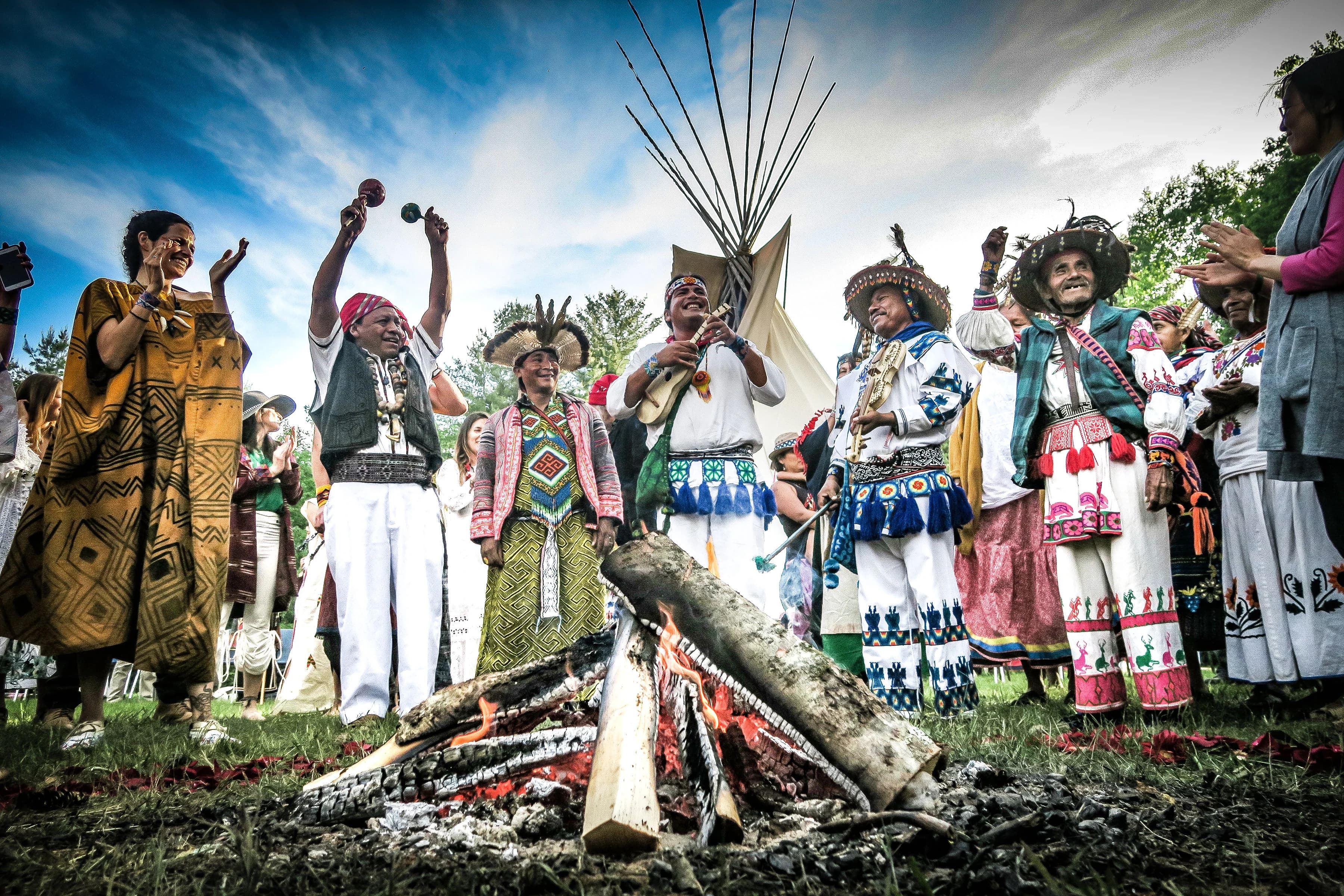 Community gathered around a ceremonial fire with a tipi under open sky