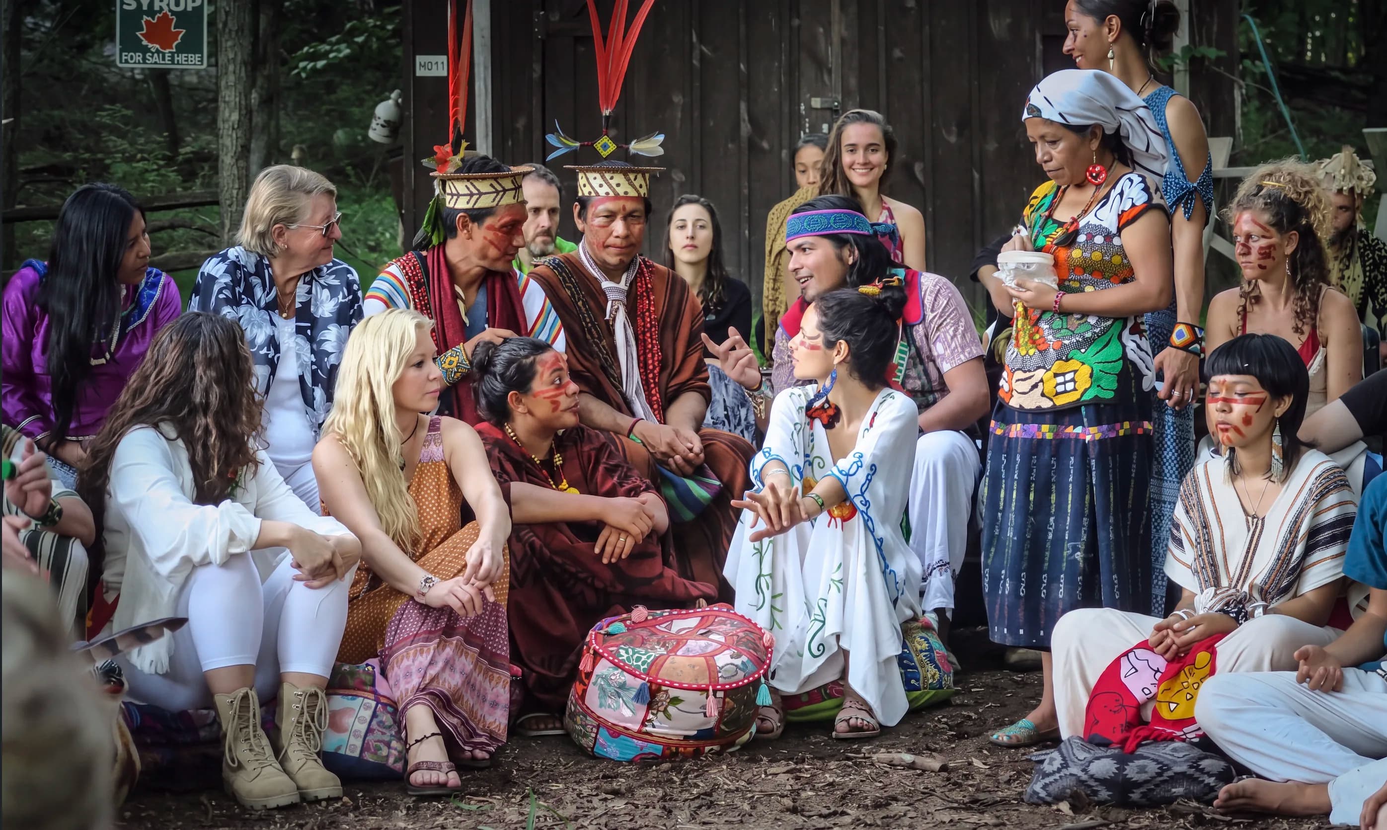 Indigenous elders and community members gathered in a circle outdoors, some in traditional ceremonial dress, engaged in conversation in a wooded setting